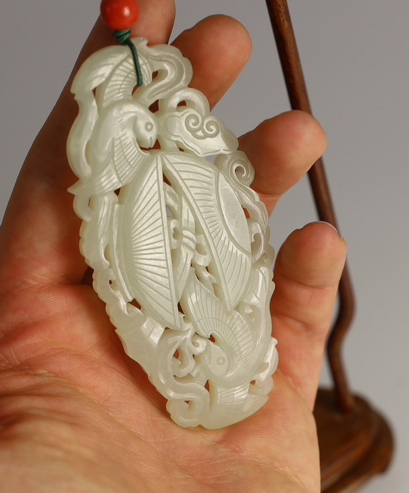 A Chinese white jade twin phoenix reticulated plaque, 19th century, 8.7cm, suspended from a wood stand with coral bead to the cord